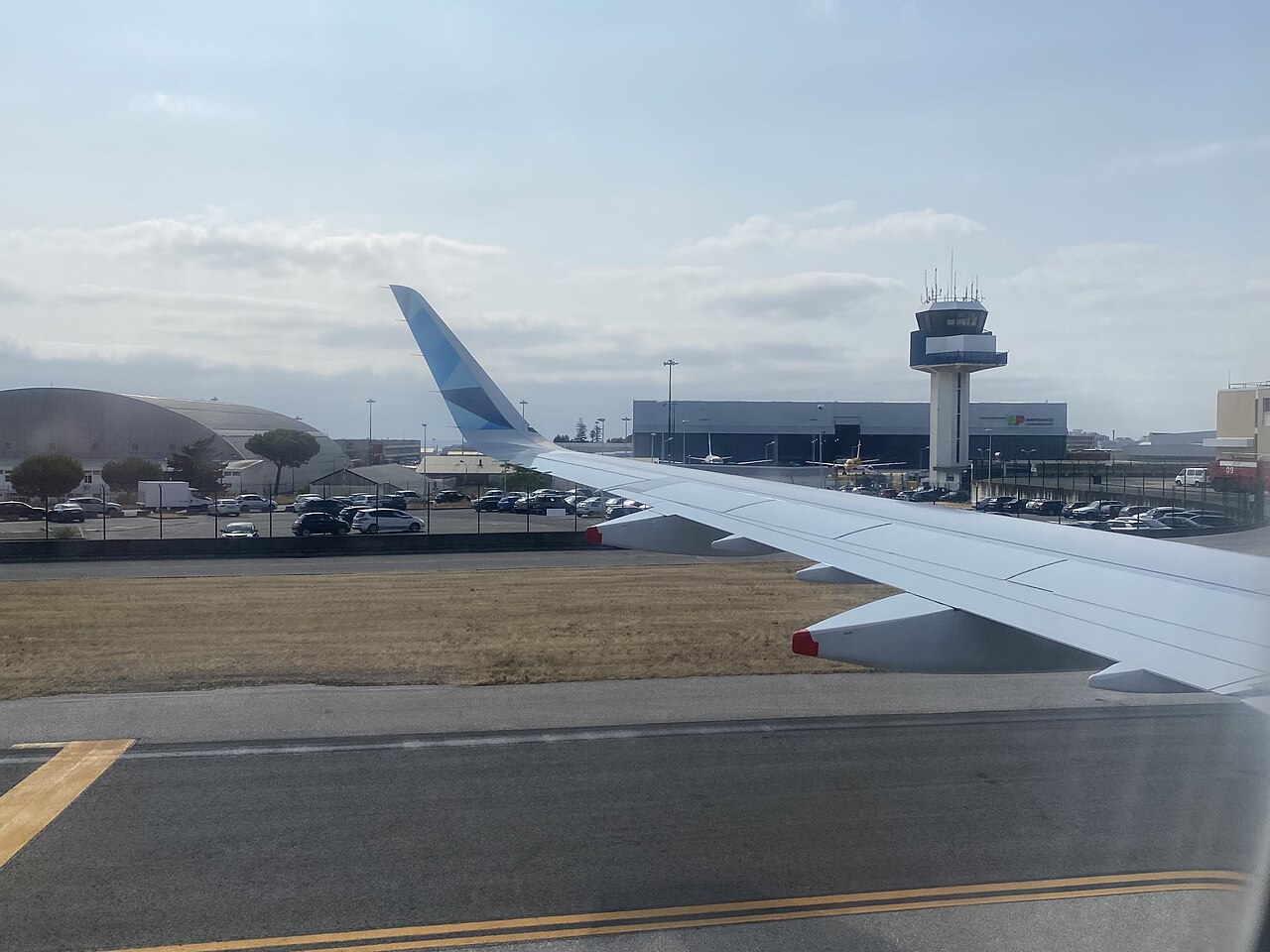 The outside view of a plane, showing Lisbon Airport's control tower. The plane wing is white with a blue tip and the sky is grey. the runway is made out of concrete.