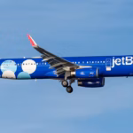 A JetBlue A321 flying under a blue sky. Its livery is mostly blue, with its tail being white, dark and light blue balls. The airport is close to landing at JFK.
