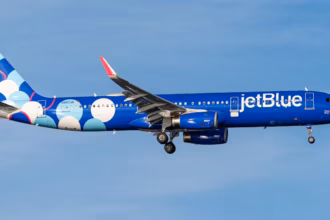 A JetBlue A321 flying under a blue sky. Its livery is mostly blue, with its tail being white, dark and light blue balls. The airport is close to landing at JFK.
