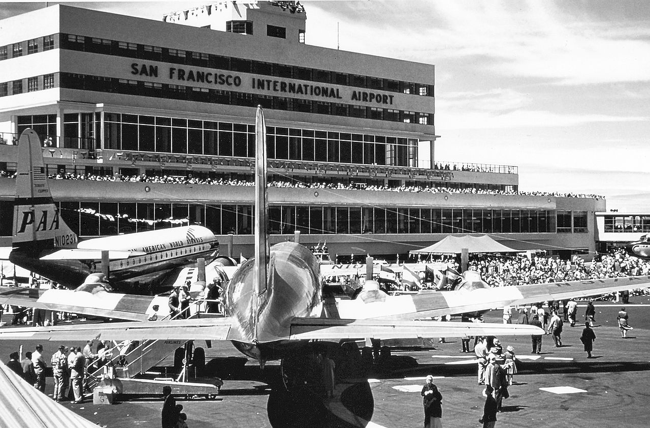 Vintage, black and white image of the SFO Terminal. The terminal is seen behind several people and aircraft.