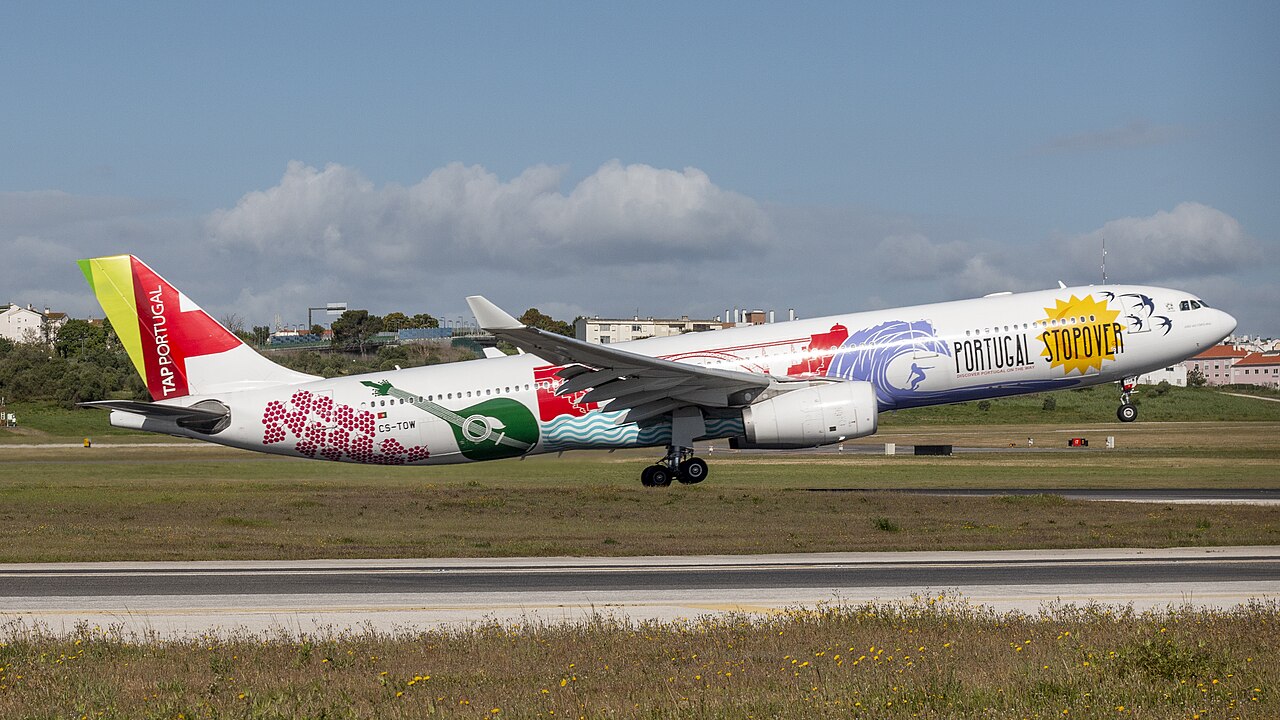 A TAP Air A330 taking off at Lisbon International Airport. The plane has a special livery displaying several Portuguese features, such as big waves, which is ver colourful. The sky is both blue and cloudy.