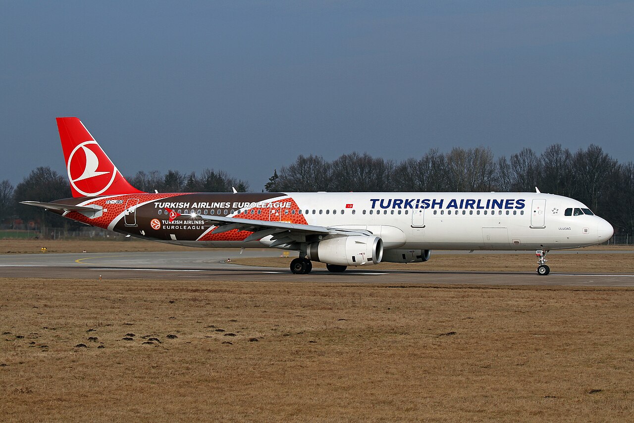 A Turkish Airlines A321-200 after landing. The aircraft displays a special livery, Euroleague, a basketball league. The aircraft is mostly white, but has its logo in blue and its mid-ends and tail are red, brown and white. The sky is grey and blueish.