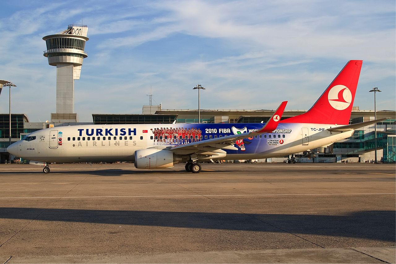 A Turkish Airlines B787-800 with a 2010 FIBA World Championship livery. The aircraft is parked in front of a control tower and terminal. The aircraft, capable of long-haul flights, is mostly white, with blue and red accents.