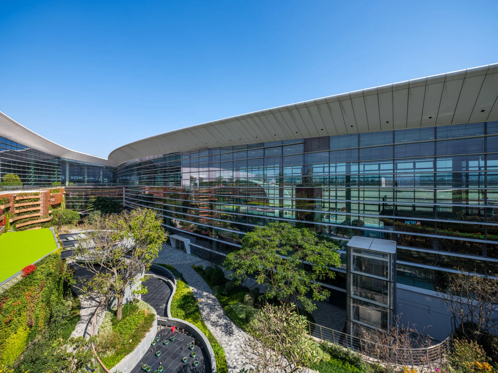 A large building with the external walls made from glass windows wraps around a beautiful natural garden scene in the centre of the image. The garden is layered at different tier levels, and boats luscious green grass, and a variety of trees flowers and foliage. The top of the image shows a clear blue sky with no clouds.