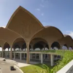 Techo airport in Cambodia is pictured from a corner angle. The roof rises and falls in curves along both sides as it extends out into the background. The roof resembles bamboo and rattan baskets. Arches can be seen all the way along both sides beneath the curved roof slopes. The background shows blue sky with minimal clouds. In the foreground grass and pathways up to the structure can be seen.