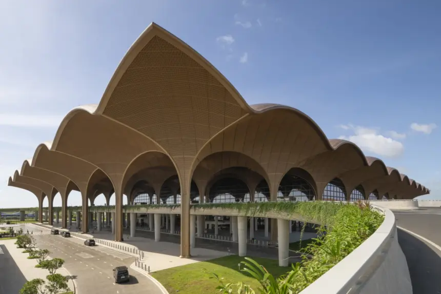 Techo airport in Cambodia is pictured from a corner angle. The roof rises and falls in curves along both sides as it extends out into the background. The roof resembles bamboo and rattan baskets. Arches can be seen all the way along both sides beneath the curved roof slopes. The background shows blue sky with minimal clouds. In the foreground grass and pathways up to the structure can be seen.