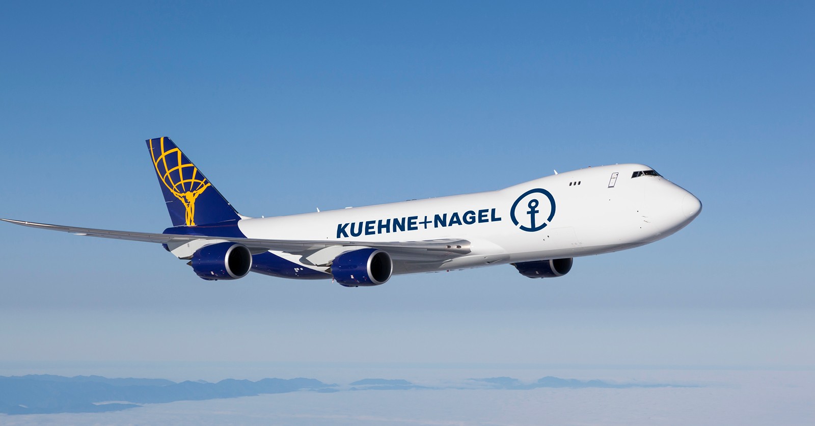 A Kuehne+Nagel aircraft flying through the blue sky. White and blue fuselage with a yellow symbol on the tail of a man holding up the earth.
