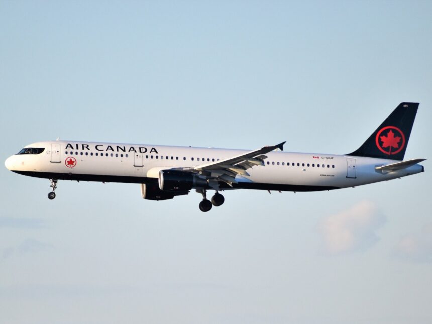A white air canada airbus A321 in flight in a light blue sky with minimal clouds. The aircraft occupied the entire centre of the image, facing left, with the nose to the left and the tail at the right. The majority of the aircraft is white, but the tail is black with the Canadian red maple leaf on it.