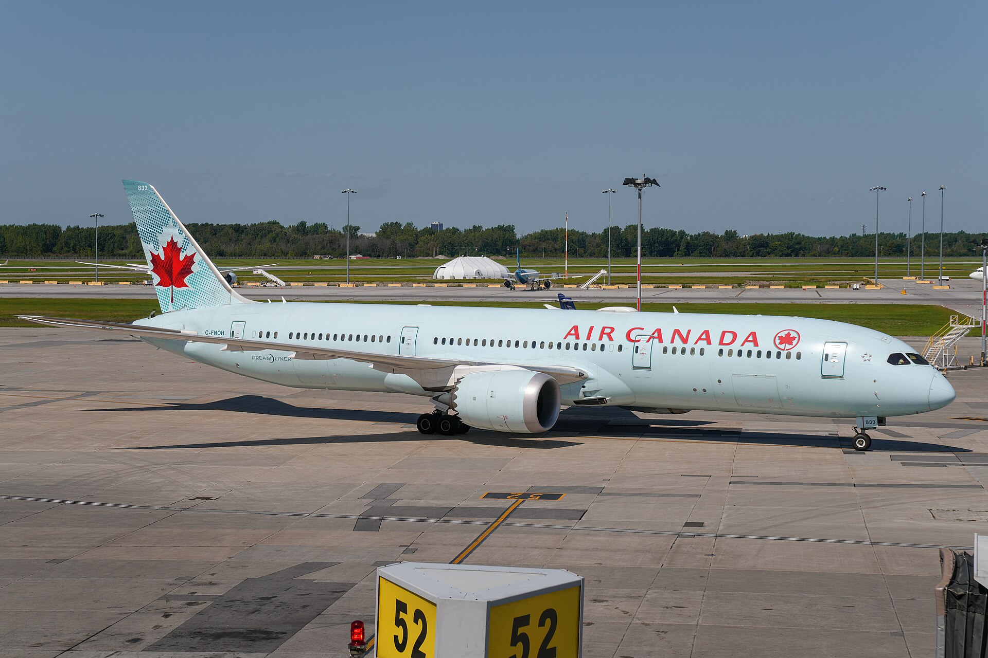 A white Air Canada Boeing 777 aircraft landed at an airport. The aircraft occupied the centre right of the image, its nose facing the right. The foreground is grey brown tarmac/concrete of an airport bay or hanger, and in the background a runway can be seen. Beyond the runway there is green grass and a row of foliage. Across the top is blue cloudless sky.