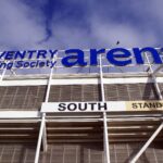 A close up image of the identifying sign on the south entrance of the Coventry Building Society Arena. The image is taking as though from below looking up. The white poles of the stadiums structure can be seen below the large cobalt blue writing. The background of the image right at the top shows a blue sky with some clouds.