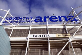 A close up image of the identifying sign on the south entrance of the Coventry Building Society Arena. The image is taking as though from below looking up. The white poles of the stadiums structure can be seen below the large cobalt blue writing. The background of the image right at the top shows a blue sky with some clouds.