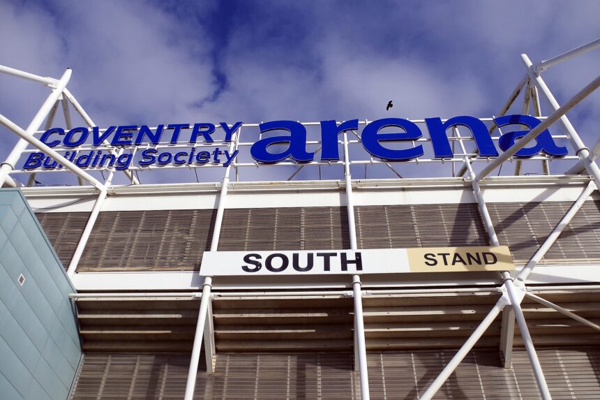 A close up image of the identifying sign on the south entrance of the Coventry Building Society Arena. The image is taking as though from below looking up. The white poles of the stadiums structure can be seen below the large cobalt blue writing. The background of the image right at the top shows a blue sky with some clouds.