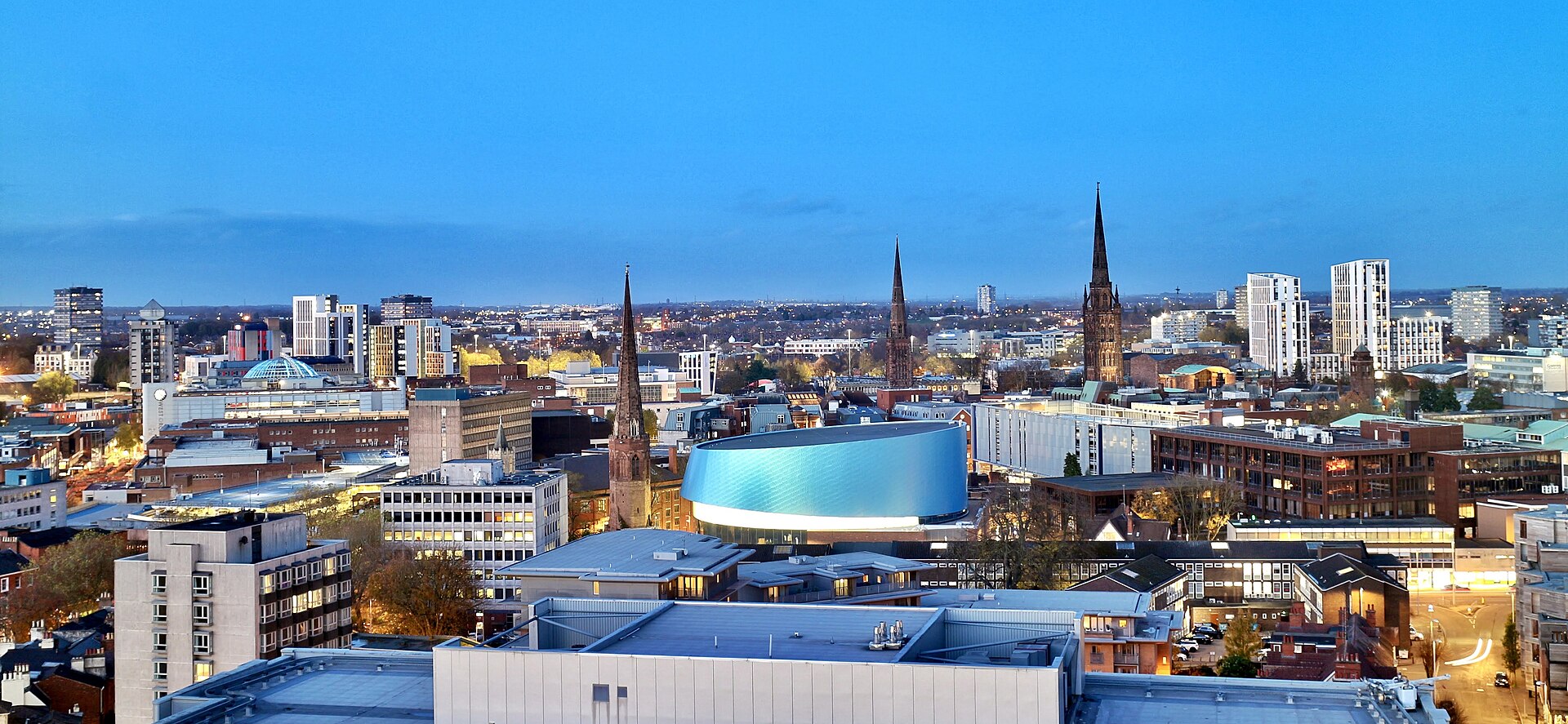 A view of Coventry skyline in the evening. Building extend out from close to the camera at the bottom of the image into the distance in the centre of the image. the top of the image depicts a darkening blue sky in what looks like early evening.