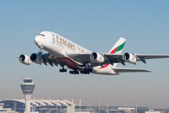 A white Emirates jet is in flight against a pale blue sky along the top background, and a cityscape along the bottom. The nose of the plan is upper centre left, the tail at lower centre right, at an angle.