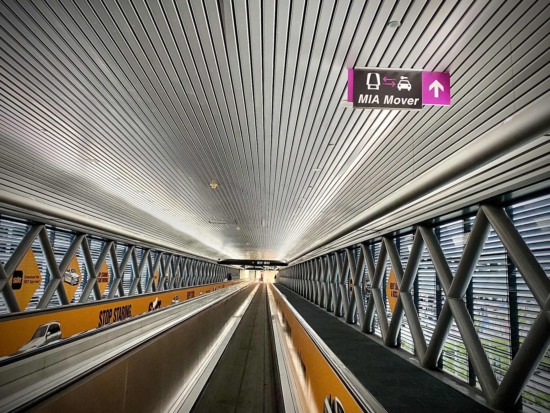 A corridor in Miami Airport with an MIA automated people mover running along the length of it. The mover is in the centre of the image, with a sign overhead on the centre right pointing to it. The walls either side have floor to ceiling windows with 'X' shaped cross pillars also all the way along.