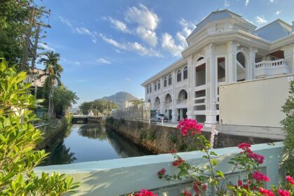 Centre right a large white domed building with ornate trim designs and archways sits of the edge of a canal, which occupies the centre left. The picture is taken from a bridge over the canal. In the foreground, flowers and foliage along the edge of the bridge can be seen. In the background another building can be seen from a distance against a blue sky with a few small clouds.