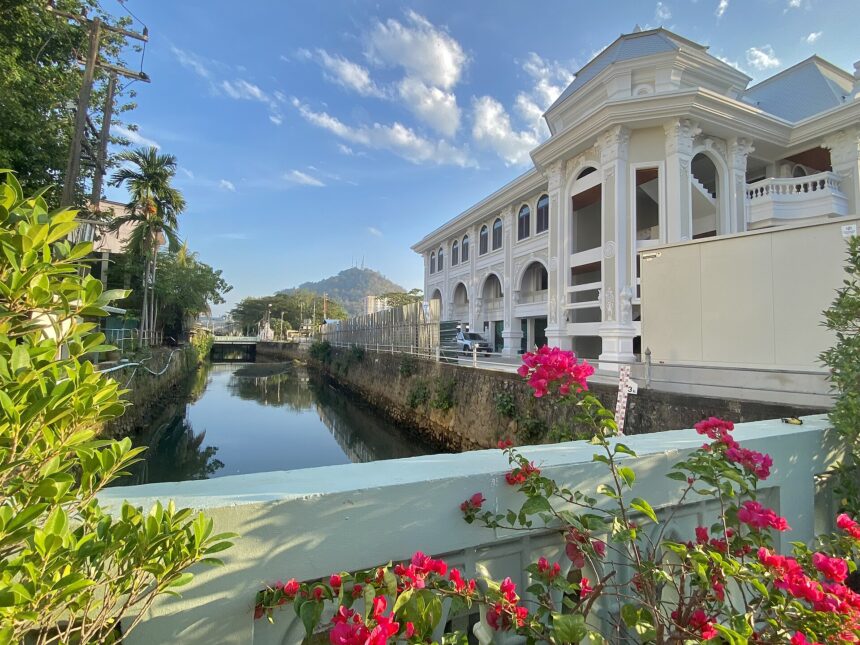 Centre right a large white domed building with ornate trim designs and archways sits of the edge of a canal, which occupies the centre left. The picture is taken from a bridge over the canal. In the foreground, flowers and foliage along the edge of the bridge can be seen. In the background another building can be seen from a distance against a blue sky with a few small clouds.