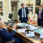Donald Trump sits in a large chair behind a desk in the Oval Office, with a map of Washington spread out on the desk, speaking and gesticulating to men in suits around the desk. A small white model of the arch is on the desk. The image is taken from slight behind Trump to his right. He is wearing a navy suit, and the other men are also all wearing suits.