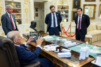 Donald Trump sits in a large chair behind a desk in the Oval Office, with a map of Washington spread out on the desk, speaking and gesticulating to men in suits around the desk. A small white model of the arch is on the desk. The image is taken from slight behind Trump to his right. He is wearing a navy suit, and the other men are also all wearing suits.