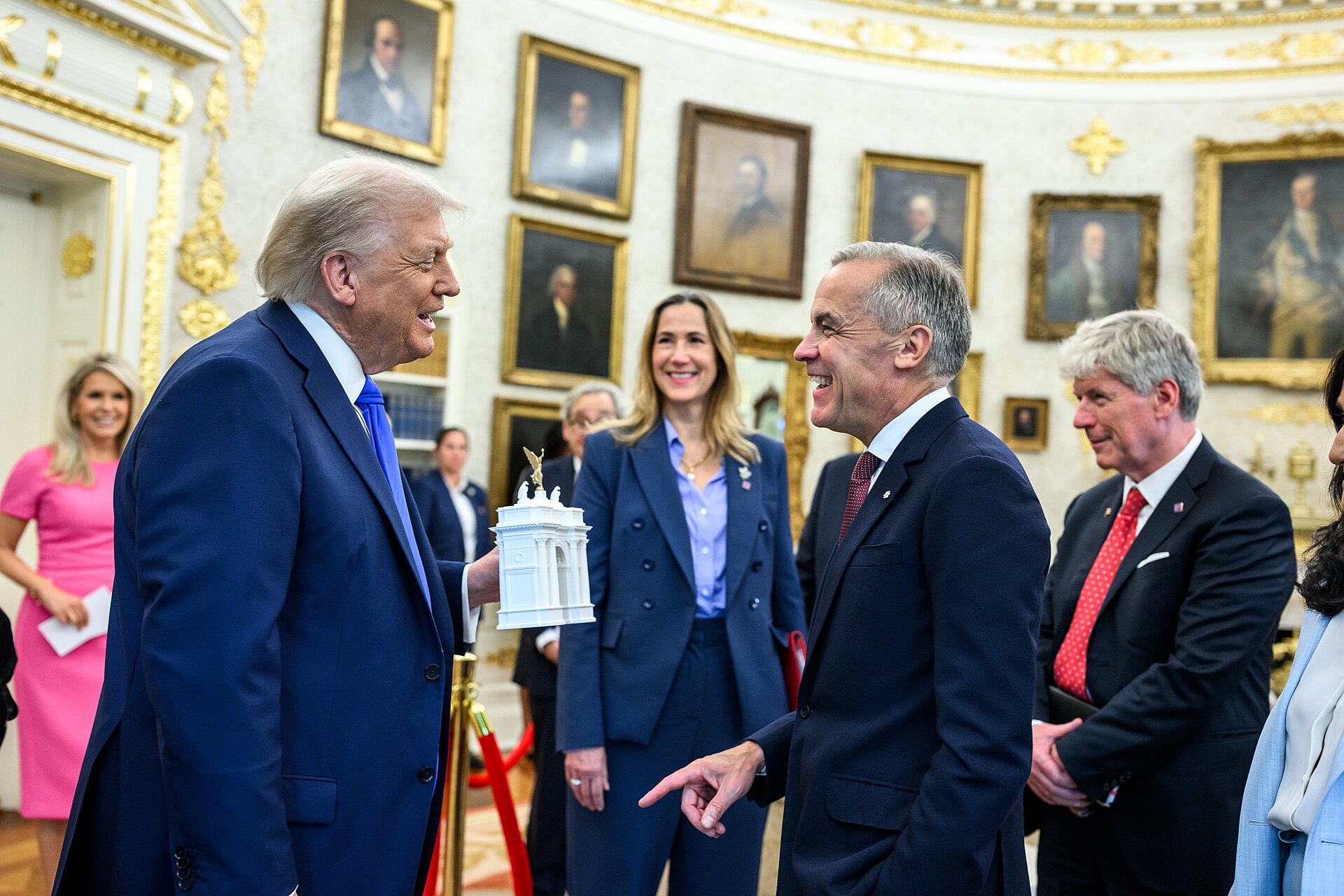 Donald Trump, on the left of the image, faces Mark Carney on the centre right, holding a small white model of his proposed arch. Trump wears a dark blue suit while Carney wears a black suit. They are both smiling and Carney is pointing slightly, and appears to be speaking. They appear to be in the White House. The background is stylistically blurred, with other individuals looking towards the two leaders. 