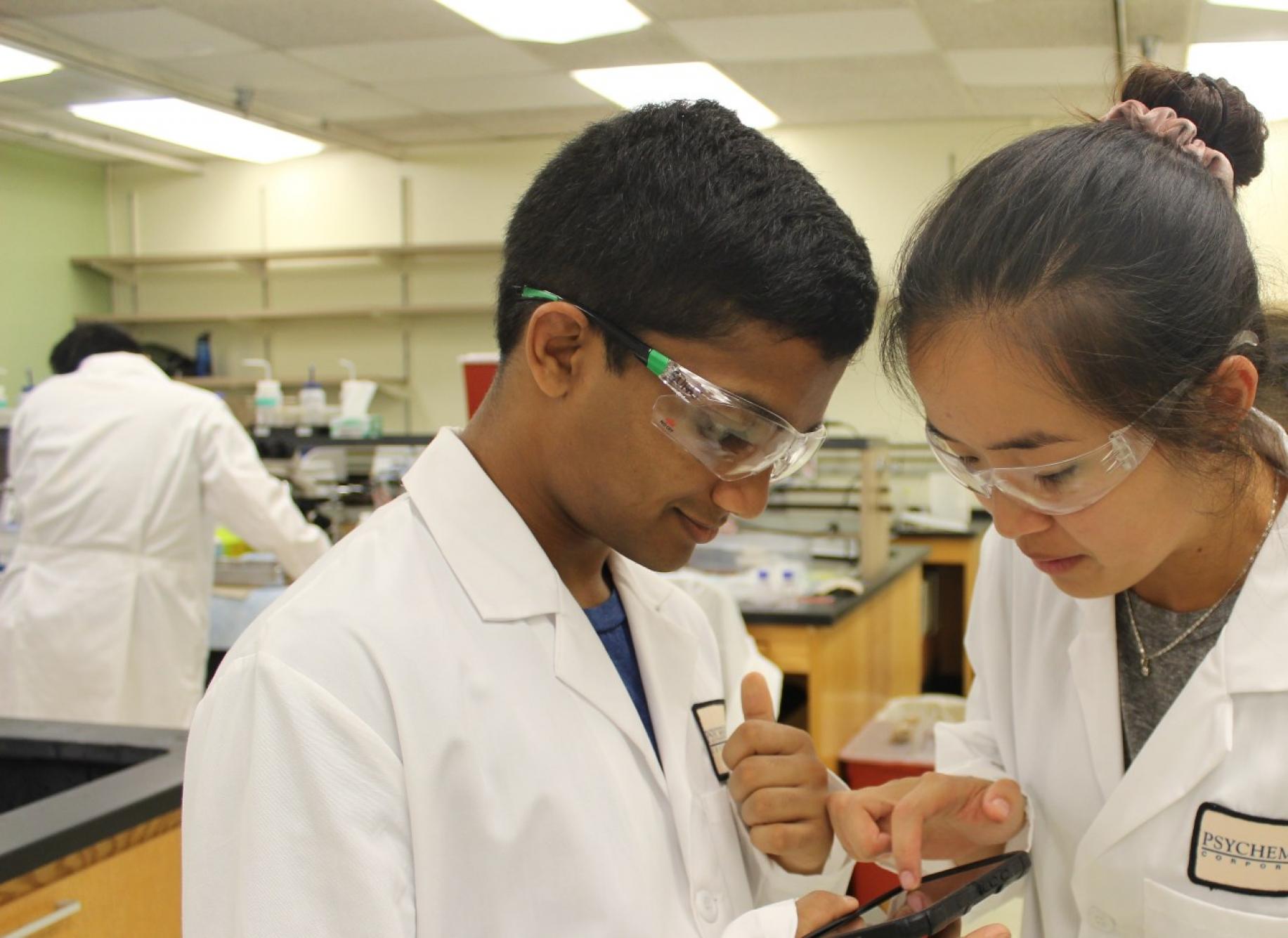 two students at the CEE in white lab jackets looking at a phone in a lab classroom environment 
