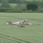 A small aircraft crash in a field, with trees in the background. The aircraft is laying upside down, no visible smoke in the photo. Red and white livery.