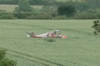 A small aircraft crash in a field, with trees in the background. The aircraft is laying upside down, no visible smoke in the photo. Red and white livery.