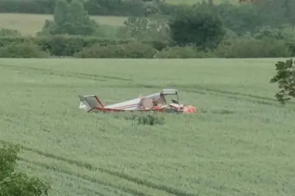 A small aircraft crash in a field, with trees in the background. The aircraft is laying upside down, no visible smoke in the photo. Red and white livery.