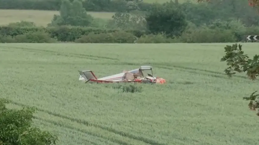 A small aircraft crash in a field, with trees in the background. The aircraft is laying upside down, no visible smoke in the photo. Red and white livery.