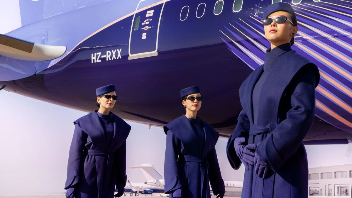 Three women standing in from of the mid-end of an aircraft. The aircraft is purple and lilac. The women are dressed in purple suits and sunglasses.