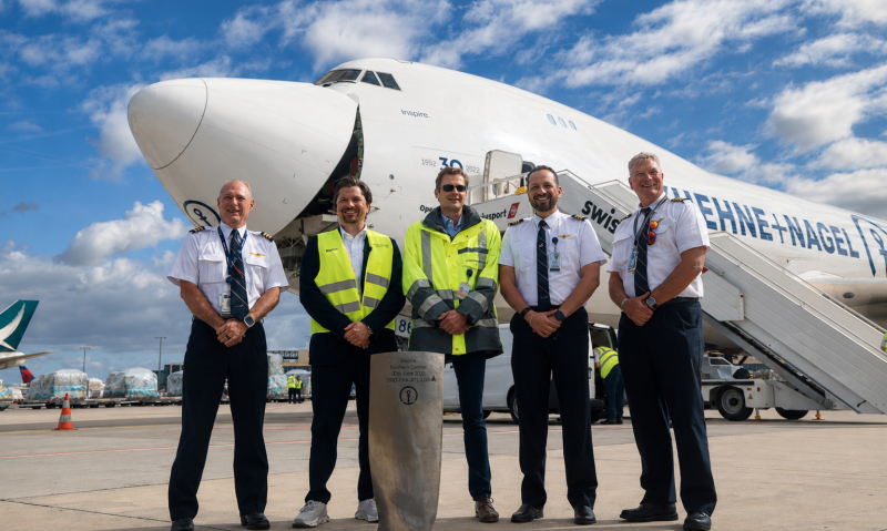 Five men stood in front of a Kuehne+Nagel aircraft on the tarmac. Cloudy, blue sky in the background. Three men in captain uniform, the other two men are in high visibility jackets.