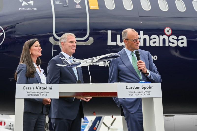 Two men and a woman stand in front of the top end of the special edition Lufthansa Airbus A320neo aircraft, behind a podium with a model of the plane on it. All three appear to be around middle aged, and two of them are named on the podium as the CEO and CTO of Lufthansa Group. The CEO holds a microphone and seems to be speaking while the other two people stand smiling. They are all dressed professionally in grey suits.