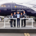 Five professionally dressed individuals from Lufthansa Group and Airbus stand holding the signed agreement, standing behind a white podium and in front of the special edition Lufthansa Airbus A320neo, which is dark blue and white. The sky is cloudy grey.