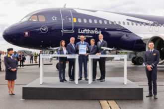 Five professionally dressed individuals from Lufthansa Group and Airbus stand holding the signed agreement, standing behind a white podium and in front of the special edition Lufthansa Airbus A320neo, which is dark blue and white. The sky is cloudy grey.