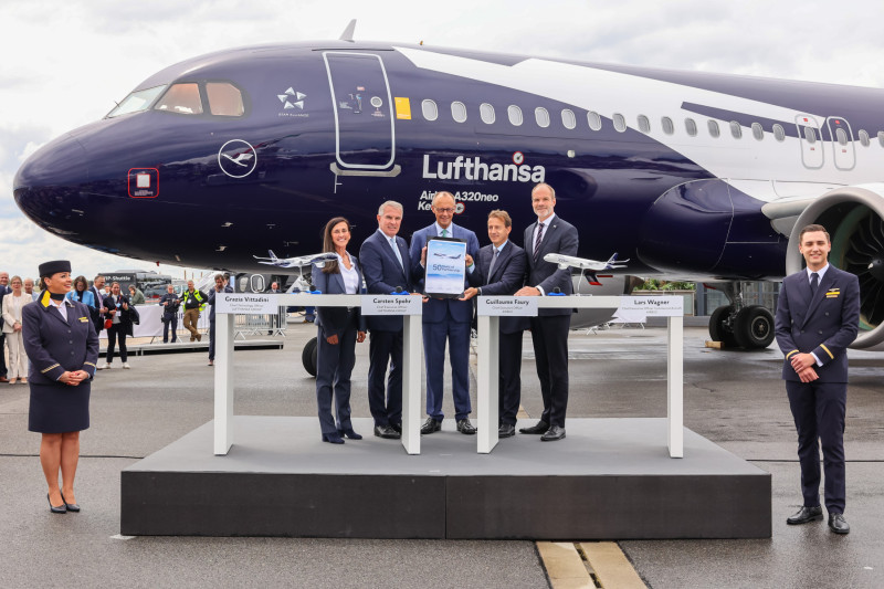 Five professionally dressed individuals from Lufthansa Group and Airbus stand holding the signed agreement, standing behind a white podium and in front of the special edition Lufthansa Airbus A320neo, which is dark blue and white. The sky is cloudy grey.