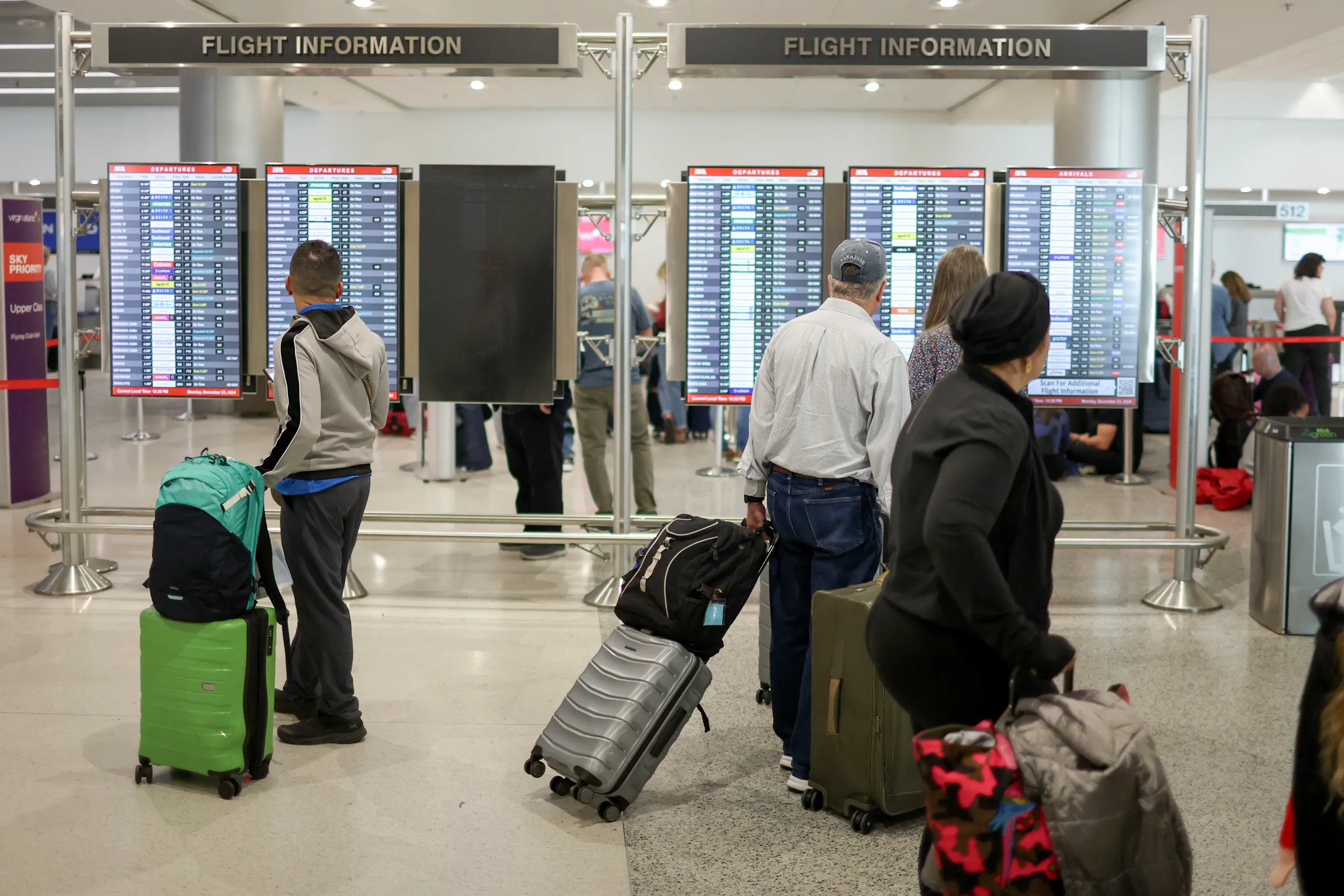 Passengers with bags and suitcases stand in front of the flight information board at Miami International Airport. Two signs at the back saying "Flight Information".