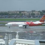 A wide, daytime shot of an Air India Express Boeing 737-800 passenger jet taxiing on a damp airport runway surface. The plane features its signature wavy red and white fuselage design and illustrative tail artwork.