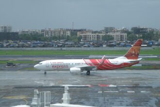 A wide, daytime shot of an Air India Express Boeing 737-800 passenger jet taxiing on a damp airport runway surface. The plane features its signature wavy red and white fuselage design and illustrative tail artwork.