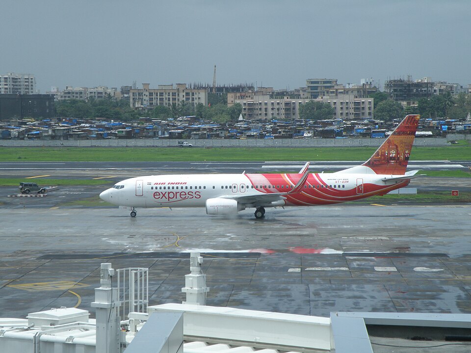 A wide, daytime shot of an Air India Express Boeing 737-800 passenger jet taxiing on a damp airport runway surface. The plane features its signature wavy red and white fuselage design and illustrative tail artwork.