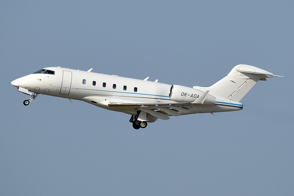 Side-profile view of a white Bombardier Challenger 300 business jet flying against a grey-blue cloudy sky. The aircraft is shown in mid-air with its landing gear extended and flaps deployed as it prepares to land