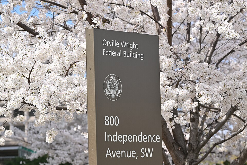 A close-up view of a dark bronze sign for the Orville Wright Federal Building displaying the address 800 Independence Avenue, SW and the Great Seal of the United States, set against a backdrop of white cherry blossom trees under a bright blue sky.