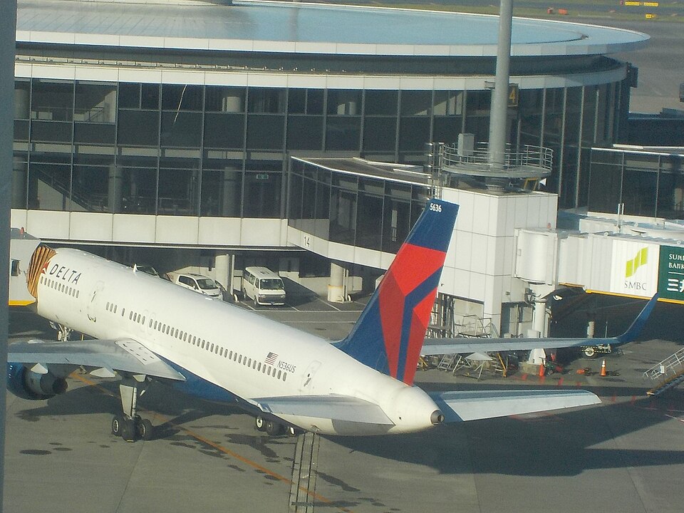 A Delta Airlines Boeing 757-200 (registration N536US) parked at an airport terminal gate with a jet bridge attached, set against a large, modern glass-walled building.