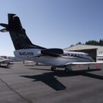 View of an Embraer Phenom 300 business jet parked on a sunlit airport apron. The aircraft features a white fuselage with a glossy black tail, engine nacelles, and upper fuselage accent
