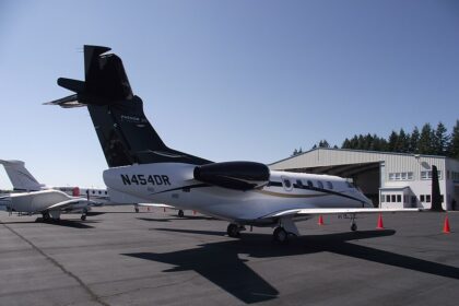 View of an Embraer Phenom 300 business jet parked on a sunlit airport apron. The aircraft features a white fuselage with a glossy black tail, engine nacelles, and upper fuselage accent