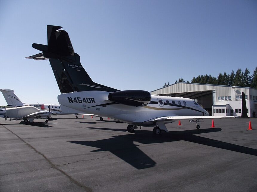 View of an Embraer Phenom 300 business jet parked on a sunlit airport apron. The aircraft features a white fuselage with a glossy black tail, engine nacelles, and upper fuselage accent