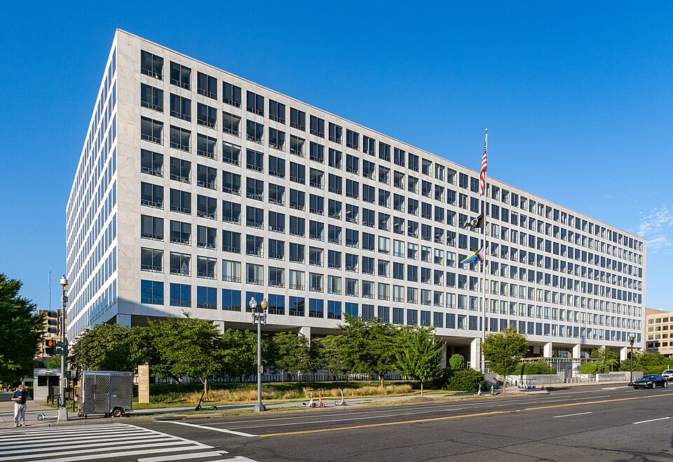 Wide exterior view of the Orville Wright Federal Building (headquarters of the U.S. Department of Transportation and Federal Aviation Administration) on a sunny day. The large, modern white concrete building with a grid of windows stands under a clear blue sky, with American and Pride flags flying on a flagpole in front.