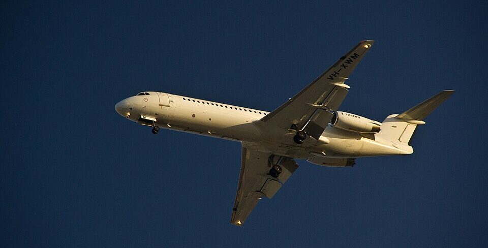A white Fokker 70 regional jet aircraft in flight against a clear deep blue sky, viewed from below with its landing gear extended. The registration 