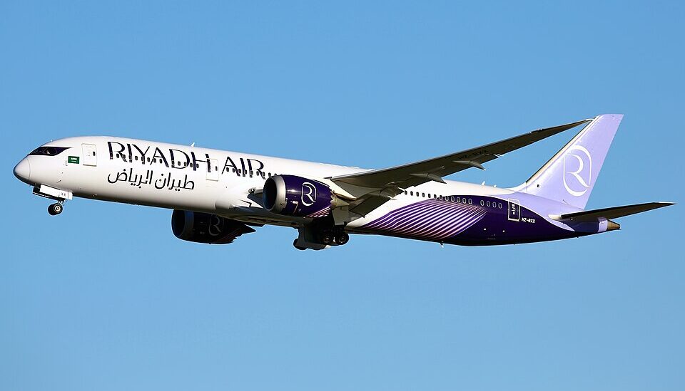 A Riyadh Air Boeing 787 Dreamliner in flight against a clear blue sky, captured in profile view with its nose landing gear extended. The aircraft displays the airline’s distinctive livery with 