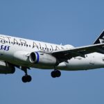 Side view of a Scandinavian Airlines (SAS) Airbus A320 in flight against a clear blue sky, captured on final approach with its landing gear fully extended. The white fuselage features “Scandinavian” written in large grey lettering along the side, with the blue “SAS” logo near the front. The engines beneath the wings are painted dark blue, and the tail is also dark blue with a white star‑shaped emblem. The aircraft’s wings are slightly flexed, with flaps deployed, and the nose is angled slightly downward as it descends.