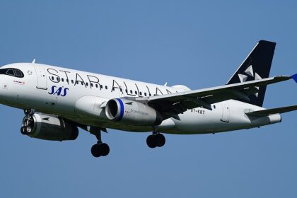 Side view of a Scandinavian Airlines (SAS) Airbus A320 in flight against a clear blue sky, captured on final approach with its landing gear fully extended. The white fuselage features “Scandinavian” written in large grey lettering along the side, with the blue “SAS” logo near the front. The engines beneath the wings are painted dark blue, and the tail is also dark blue with a white star‑shaped emblem. The aircraft’s wings are slightly flexed, with flaps deployed, and the nose is angled slightly downward as it descends.
